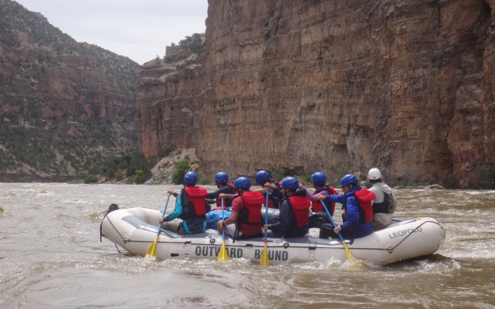 A group of people wearing safety gear paddle a raft. The river is framed by tall canyon walls. 